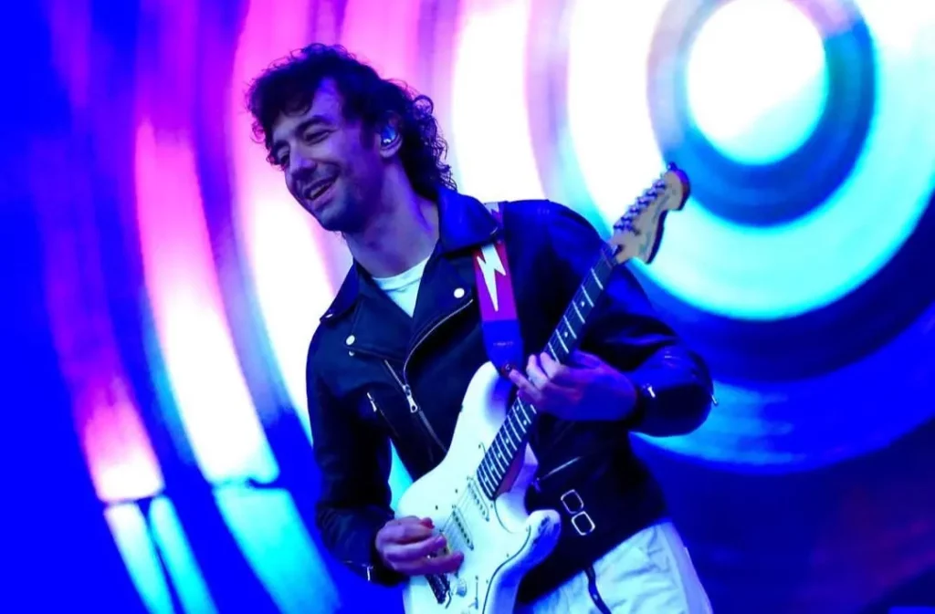 Foto colorida de Albert Hammond Jr tocando guitarra, sorrindo para o público. Ele usa jaqueta de couro preta sobre roupas brancas e a iluminação do palco é azul arroxeado com uma espiral ao fundo, como um alvo.