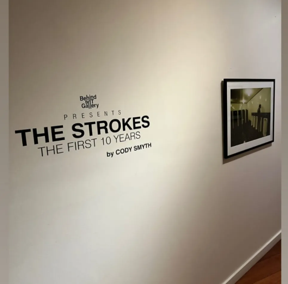 Foto colorida de parede bege com os dizeres Behind The gallery The Strokes the first ten years by Cody Smyth em letras pretas, ao lado de fotografia emoldurada.
