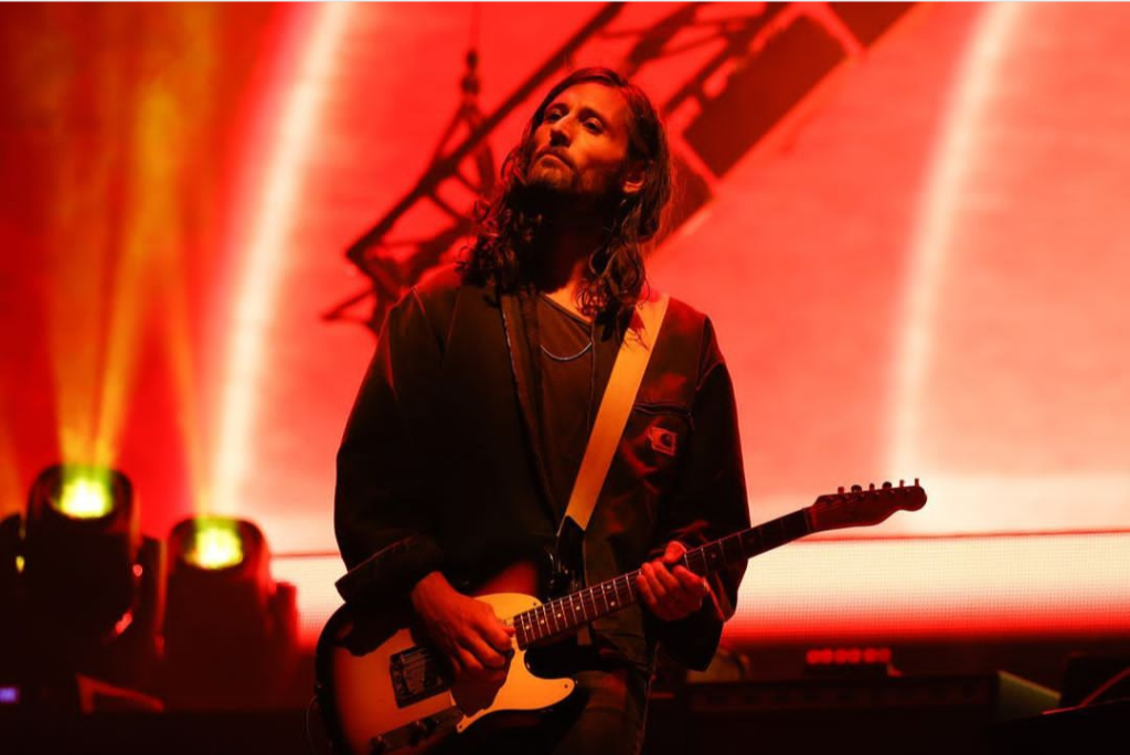 Foto colorida de Nick tocando guitarra em show, usando camisa de manga comprida sobre camiseta, ambas pretas.  A foto tem uma iluminação vermelho alaranjada que tinge quase tudo. Seu queixo está erguido e os lábios cerrados.