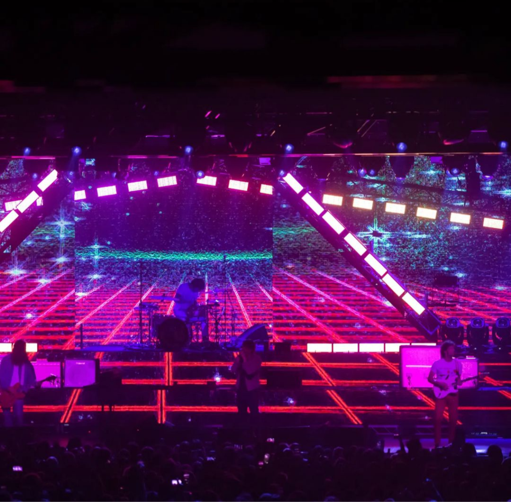 Foto colorida de palco, à distância. A iluminação tem tons de rosa claro e rosa neon que faz o piso do palco parecer quadriculado. Ao fundo, tingidos de rosa e azul, percebemos Nick Valensi, Julian Casablancas e Fabrizio Moretti tocando sua bateria.