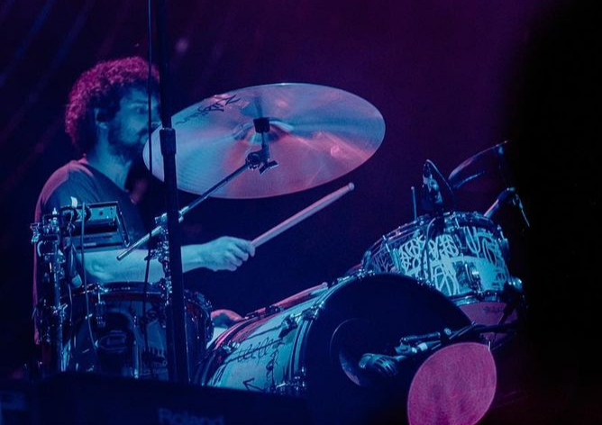 Foto colorida de Fabrizio tocando bateria em show à noite. Os tons de luz da foto são azul e roxo. Ele usa camiseta escura de manga curta, seu rosto parcialmente coberto por um dos pratos da bateria.